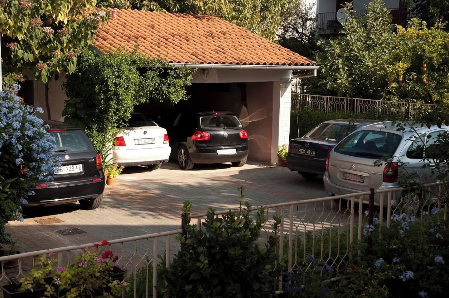 A residential garage with three cars parked inside, surrounded by greenery and a metal fence.