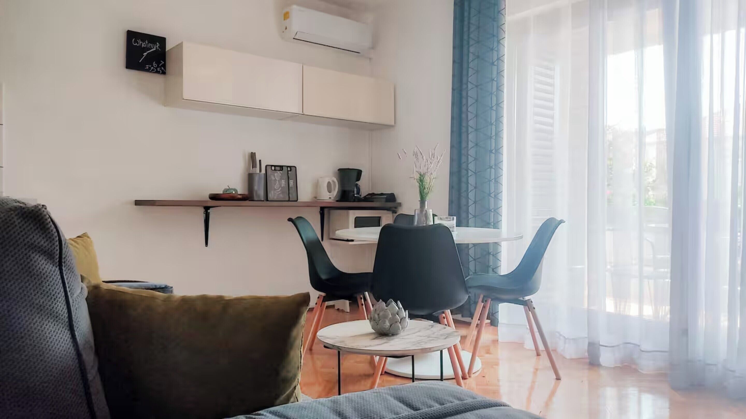 A modern living room with a sofa, a coffee table, and a dining area with chairs and a small table. There is a shelf with various items and a window with curtains.