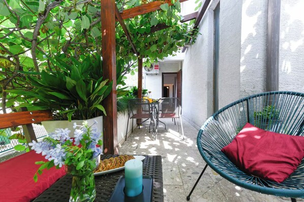 Área de pátio com pérgola de madeira, cadeira de vime com almofada vermelha, mesa com uma vela e vista para a entrada do hotel.