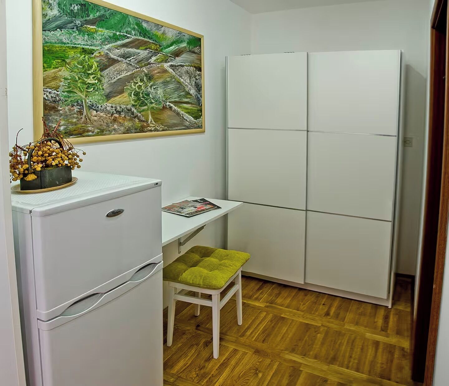 A compact kitchenette with a refrigerator, a small table, a bench, and a painting on the wall.