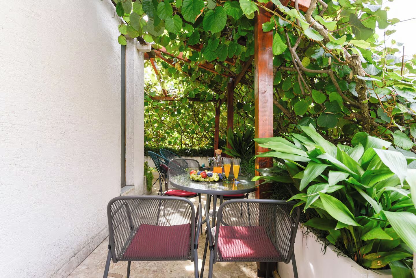 An outdoor patio with a round table and two chairs, surrounded by lush greenery and a wooden pergola.