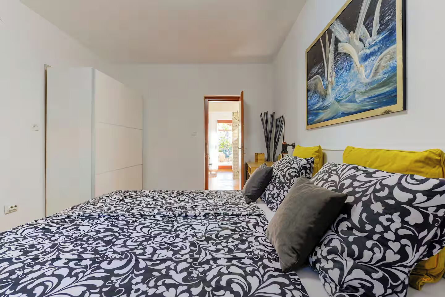 A bedroom with a bed, a painting, and a view of the outside.