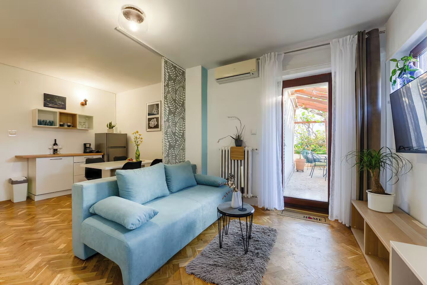 A modern living room with a light blue sofa, a dining area with a kitchen in the background, and a view of the outdoors through a glass door.