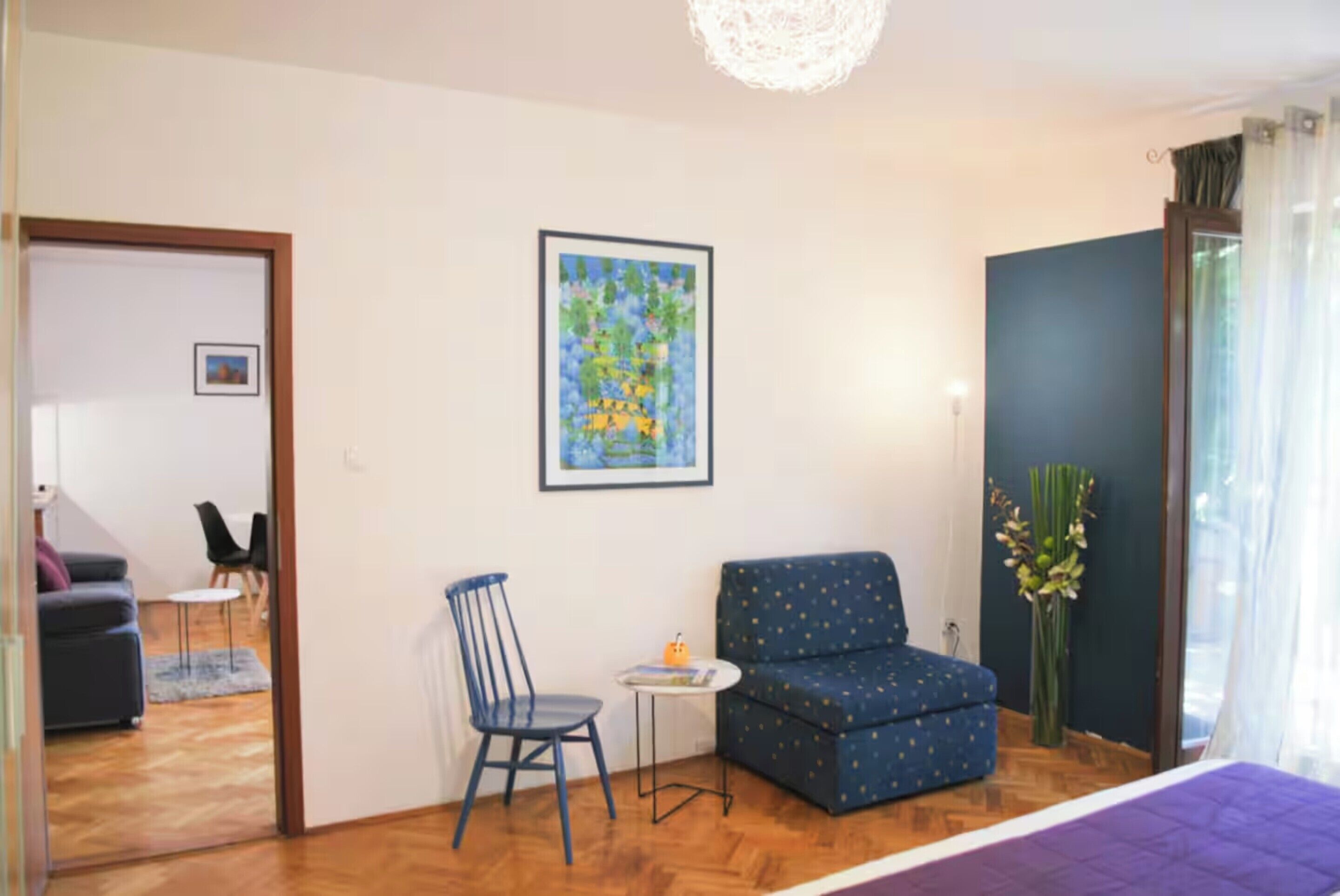 A room with a blue sofa, a small table, a chair, a framed artwork, and a window with curtains.