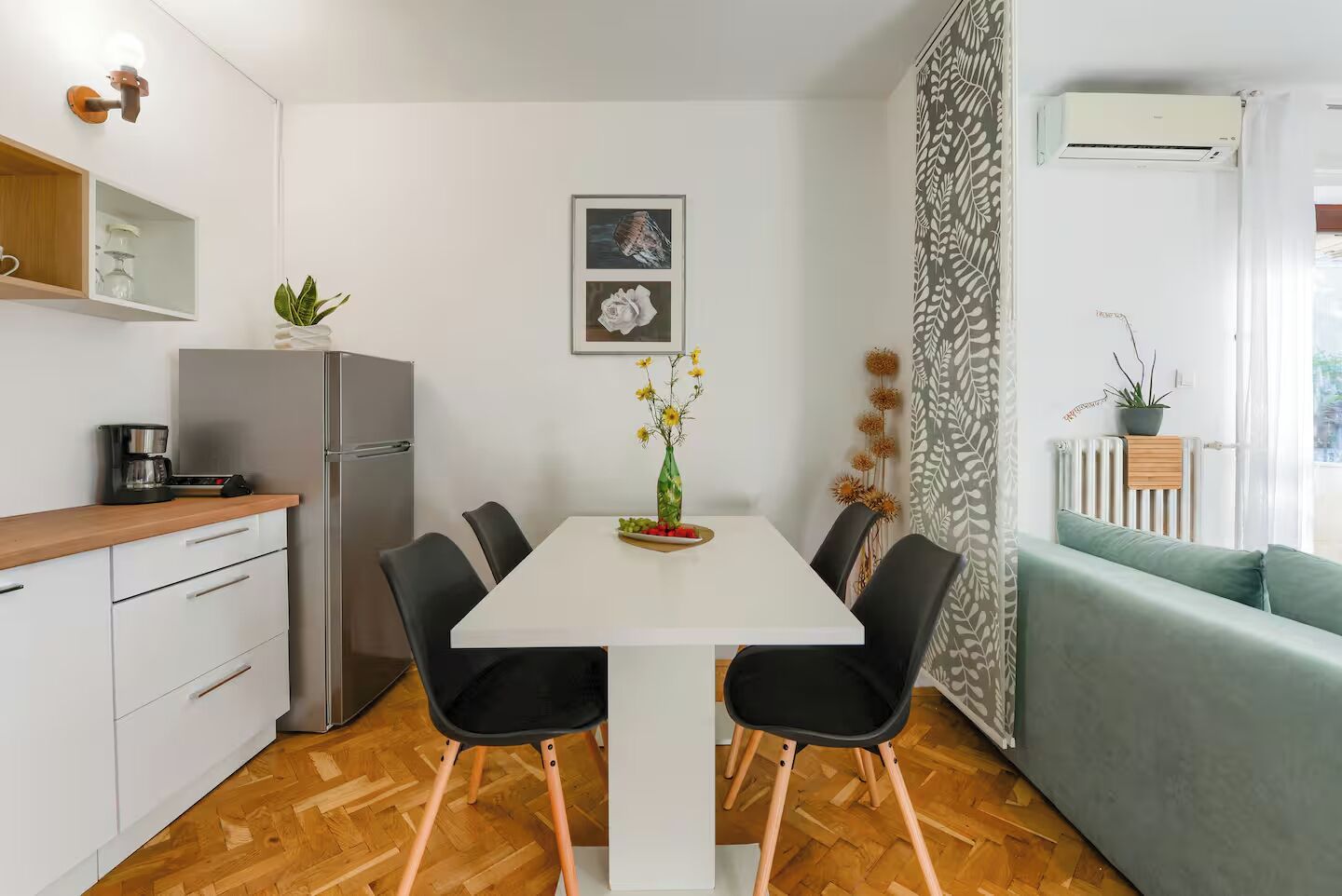 A modern kitchen with a white table, black chairs, and a refrigerator. There is a plant on the counter and a framed picture on the wall.