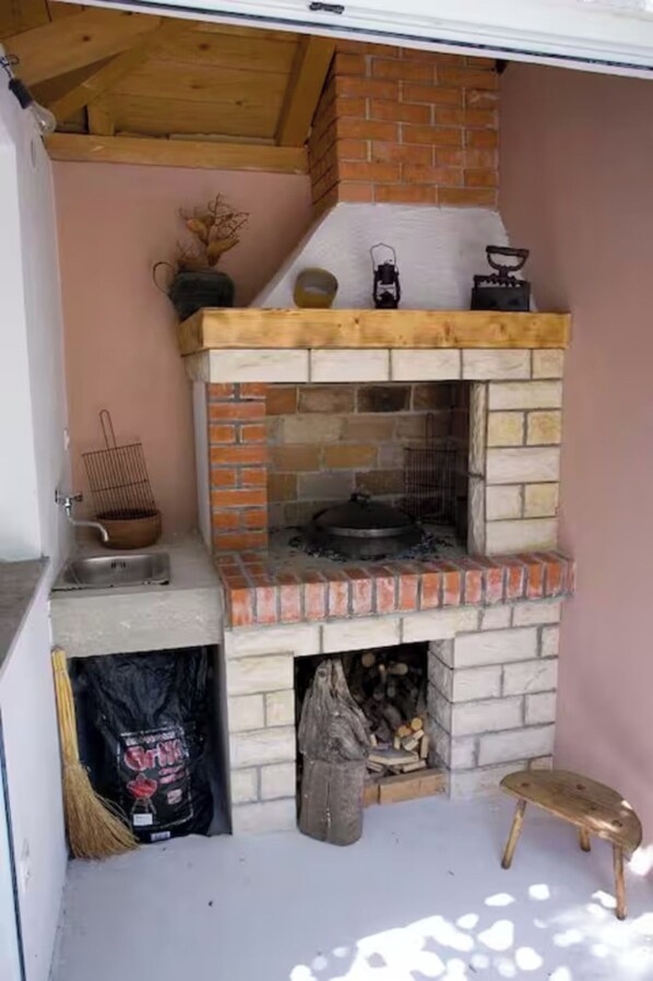 Egy téglából épített, kültéri sütő, melynek teteje fából készült, van rajta egy akasztós edénytartó, és mellette egy zsák tűzifa.
