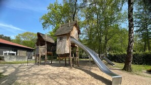 A playground with a slide, climbing structures, and a wooden house. - Chalet on Park 't Veluws Hof in Hoenderloo - With Shared Swimming Pool in Summer Season (Hoenderloo)