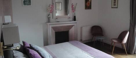 A bedroom with a bed, a chair, a fireplace, and a mirror.
