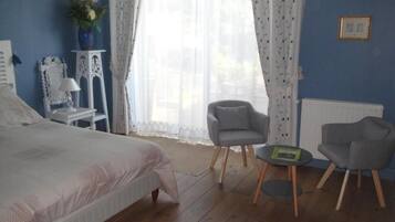 A bedroom with a bed, two grey armchairs, a small table, a white lamp, a vase with flowers, and a window with sheer curtains.