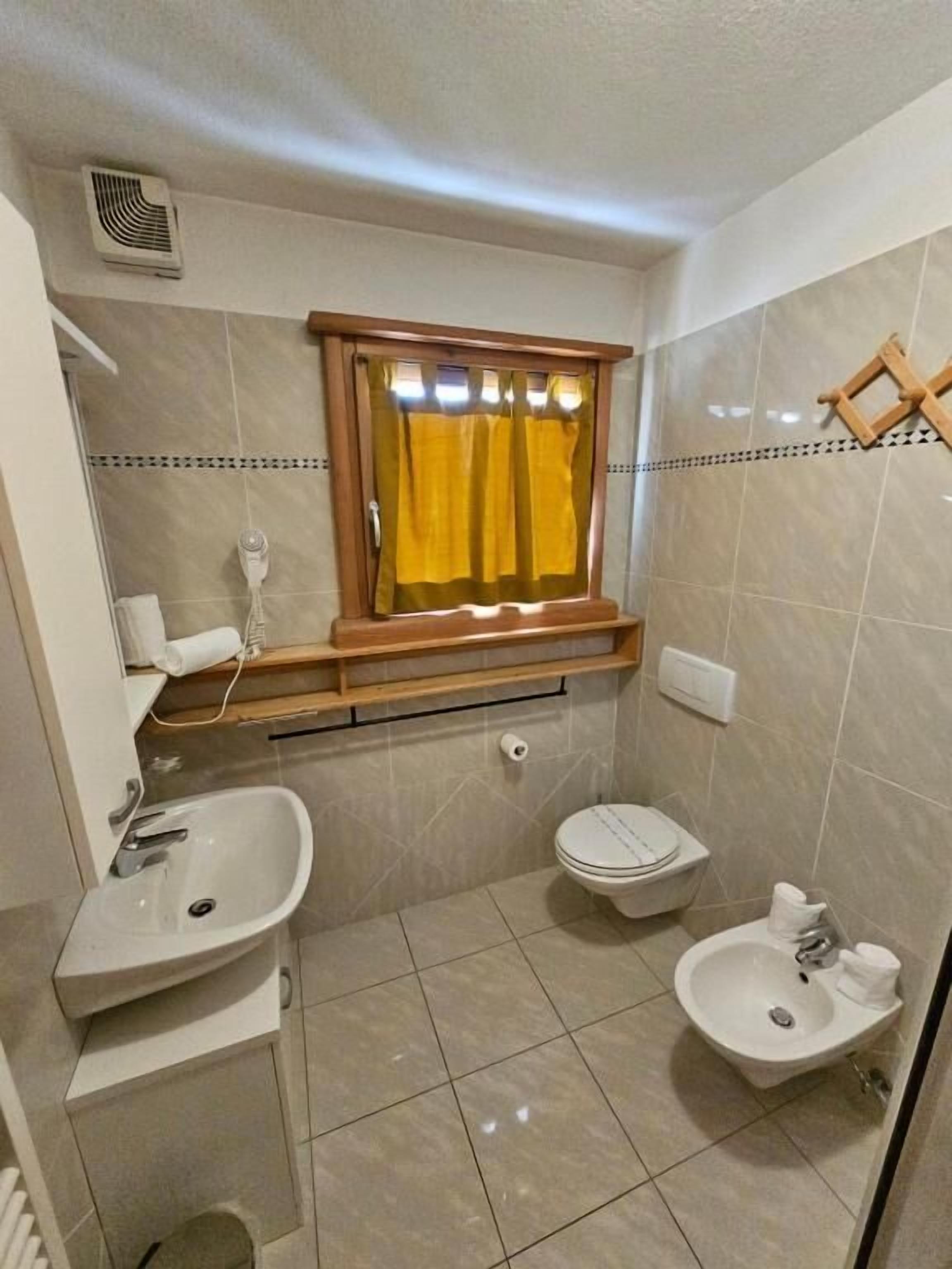 Une petite salle de bain avec un lavabo, des toilettes et un bidet. Il y a une fenêtre avec un rideau jaune et une étagère en bois au-dessus du lavabo.