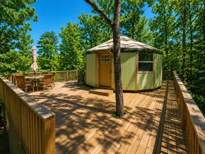 A wooden deck with a dining area, a small building with a door and window, surrounded by trees.