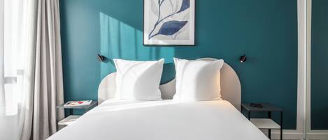Ein Hotelzimmer mit Bett, weißen Kissen, einem gerahmten Kunstwerk an der Wand, einem Nachttisch mit Lampe und einem Fenster mit Vorhängen.