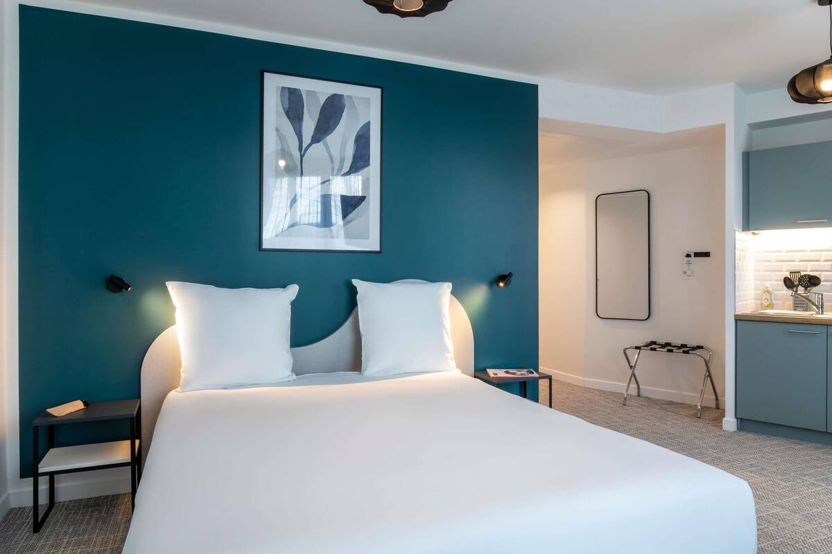 Ein modernes Hotelzimmer mit einem großen Bett, einem Nachttisch, einer Wandleuchte, einem gerahmten abstrakten Kunstwerk und einer Küchenzeile.