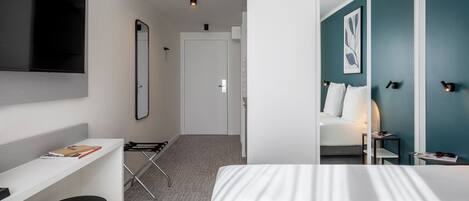 Habitación de hotel con cama, escritorio, silla, televisor montado en la pared y puerta que da a otra habitación.