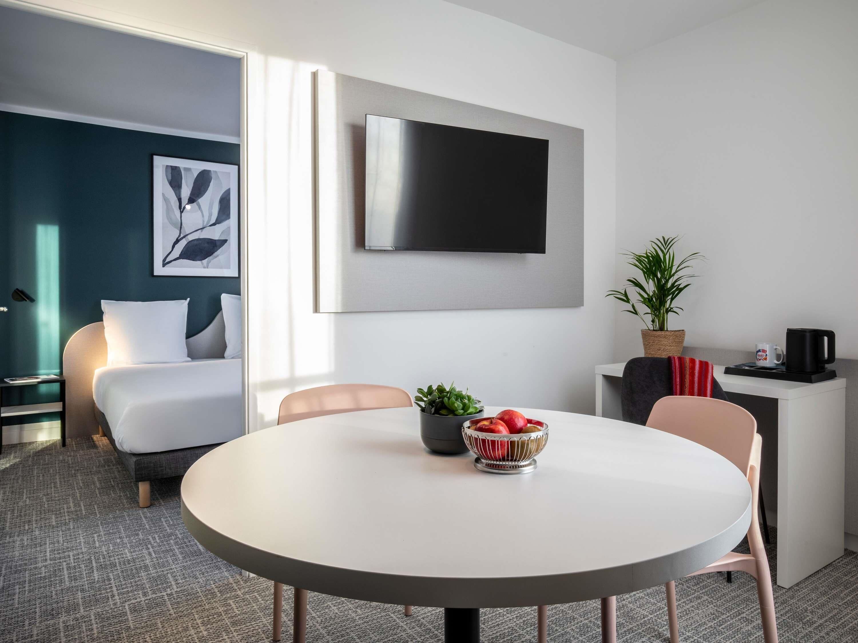 A modern hotel room with a bed, a round dining table with chairs, a TV mounted on the wall, a potted plant, and a coffee maker.