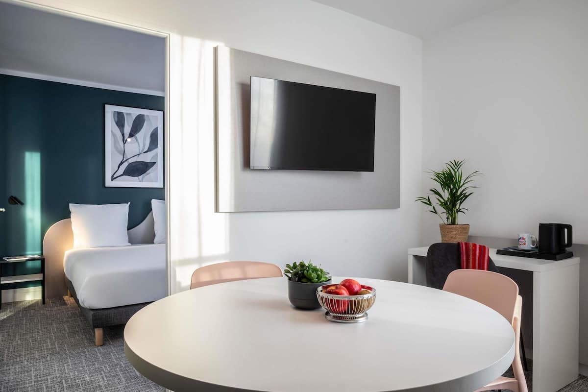 Una habitación de hotel moderna con cama, una mesa redonda con sillas, televisor montado en la pared, una planta en maceta y una cafetera.
