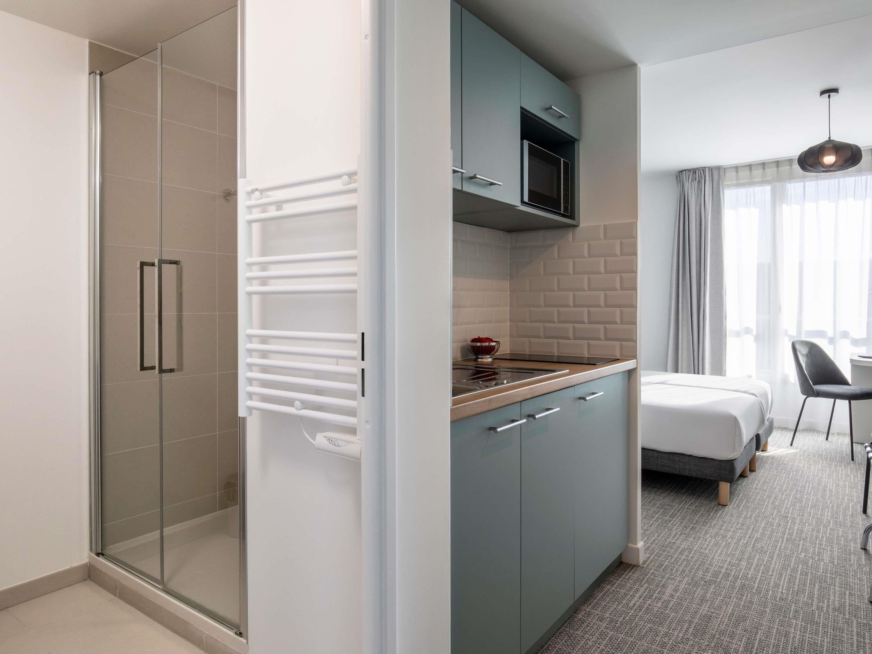 A modern hotel room with a glass shower enclosure, a kitchenette with a microwave, and a bed with a grey headboard.