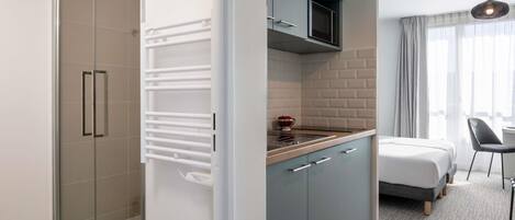 A modern hotel room with a glass shower enclosure, a kitchenette with a microwave, and a bed with a grey headboard.