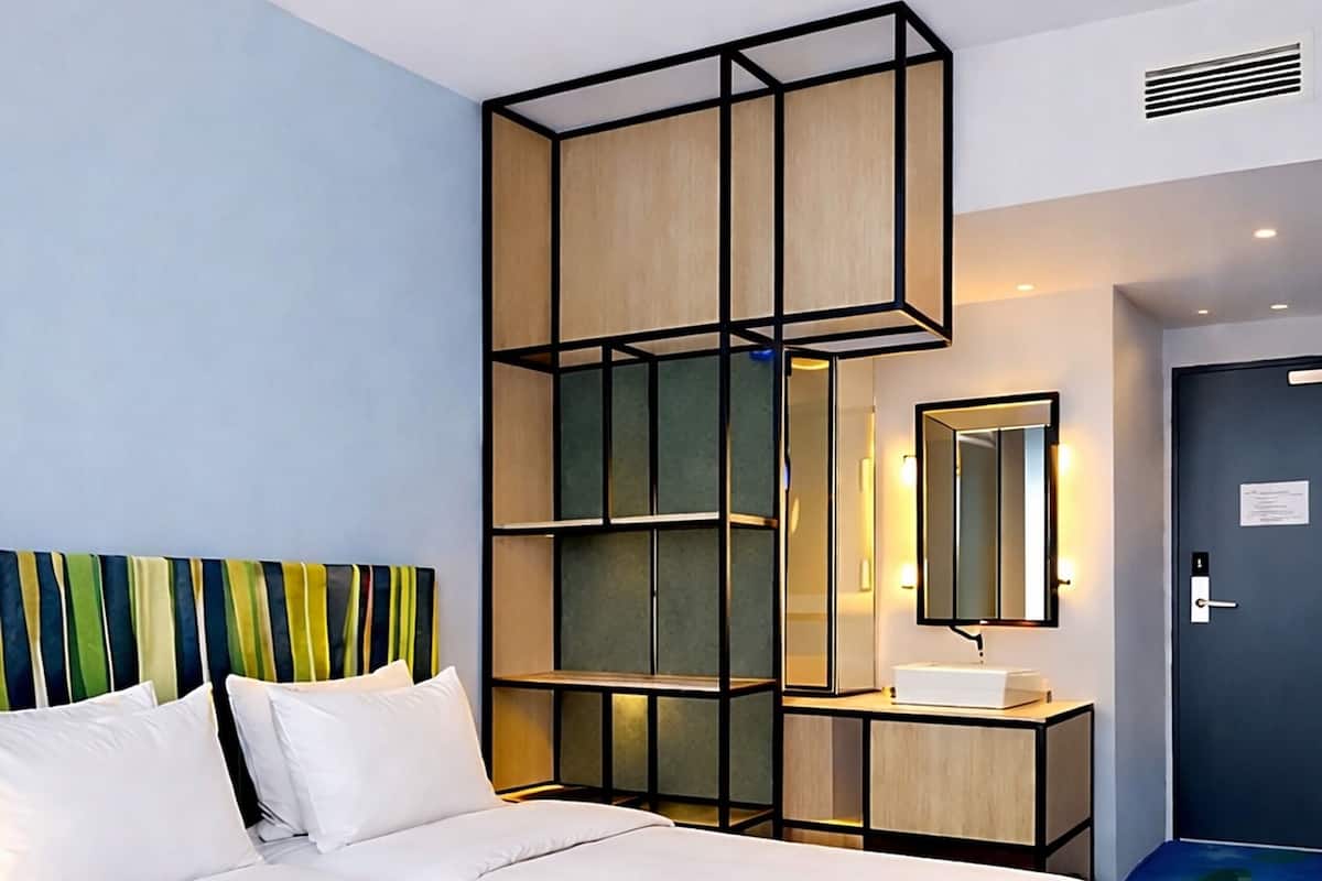 Habitación de hotel con una cama grande, cabecera de madera, mesita de noche con espejo y alfombra azul estampada.