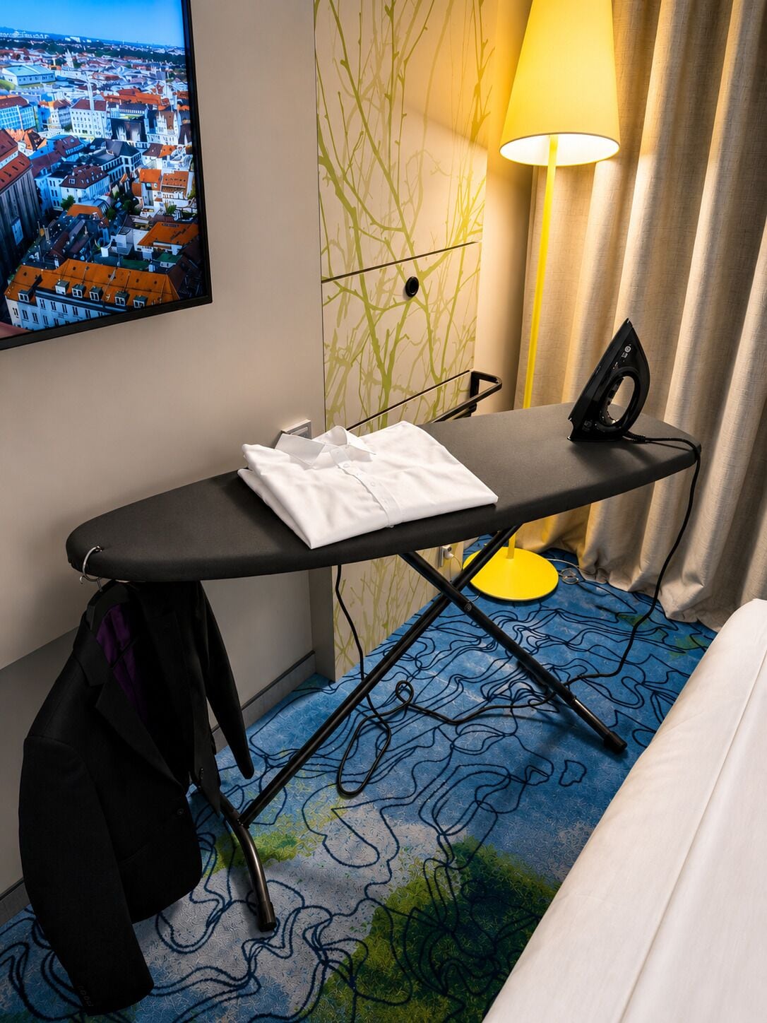 A neatly made bed, an ironing board with a white shirt, an iron, a coat hanging, a lamp, and a wall-mounted TV displaying a cityscape.