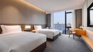 A hotel room with two beds, a desk, a chair, and a large window offering a city view.