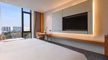 Una habitación de hotel moderna con una cama grande, un escritorio con silla, un televisor montado en la pared y vistas de la ciudad a través de amplias ventanas.