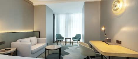 A modern hotel room with a sofa, two chairs, a desk, and a lamp.