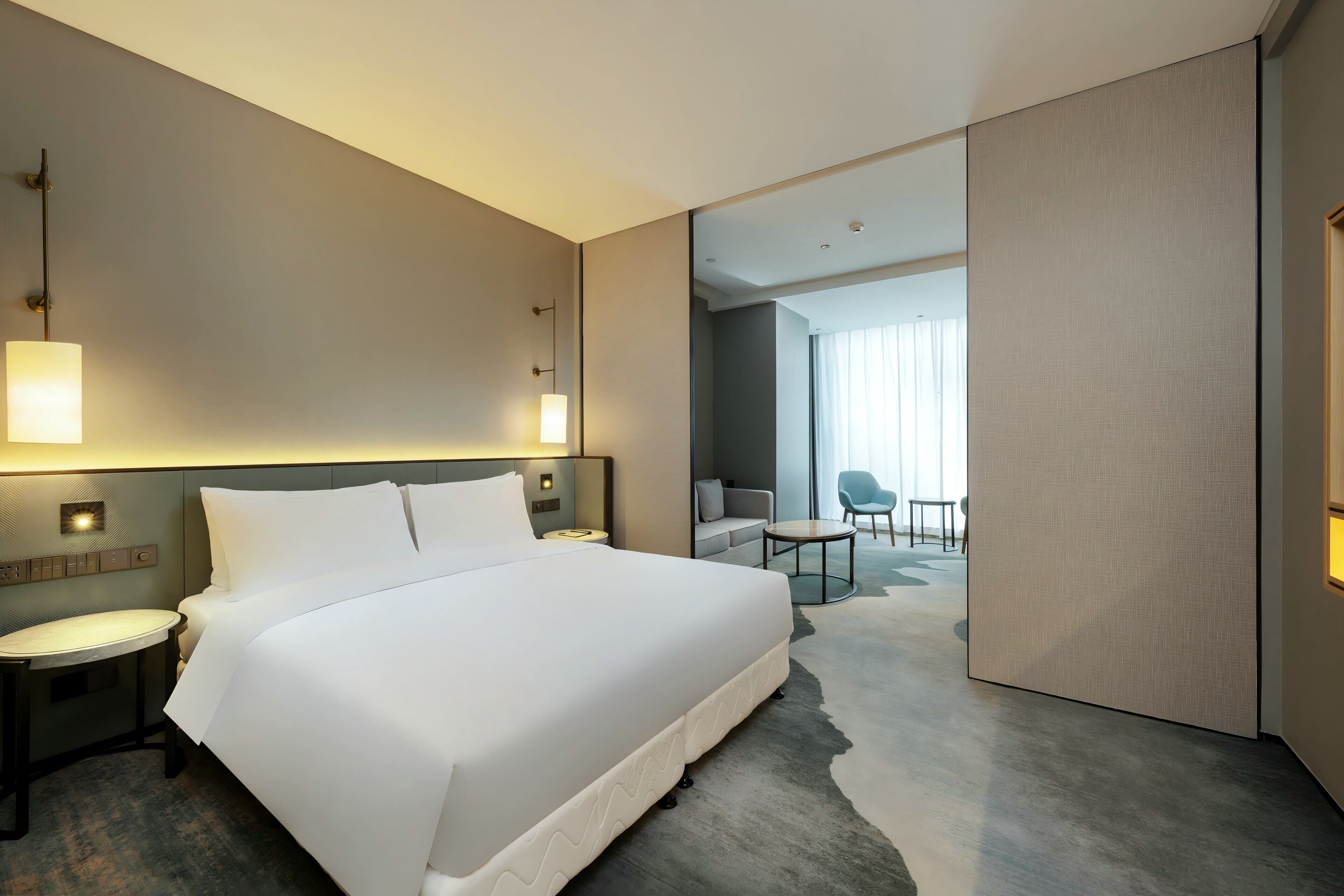 Quarto de hotel moderno com uma cama grande, mesas de cabeceira, uma escrivaninha e vista para o quarto vizinho.