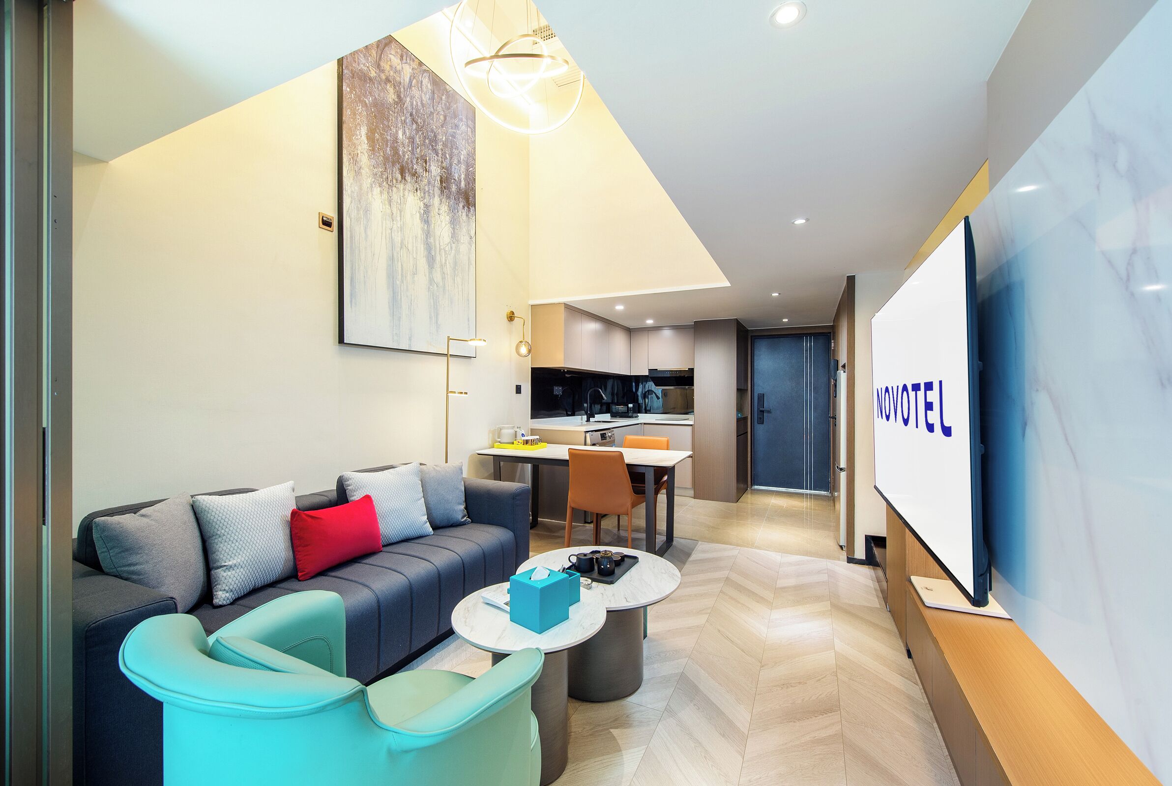 A modern hotel room with a sofa, a coffee table, a dining area, and a kitchen in the background.