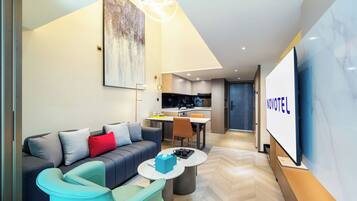 A modern hotel room with a sofa, a coffee table, a dining area, and a kitchen in the background.