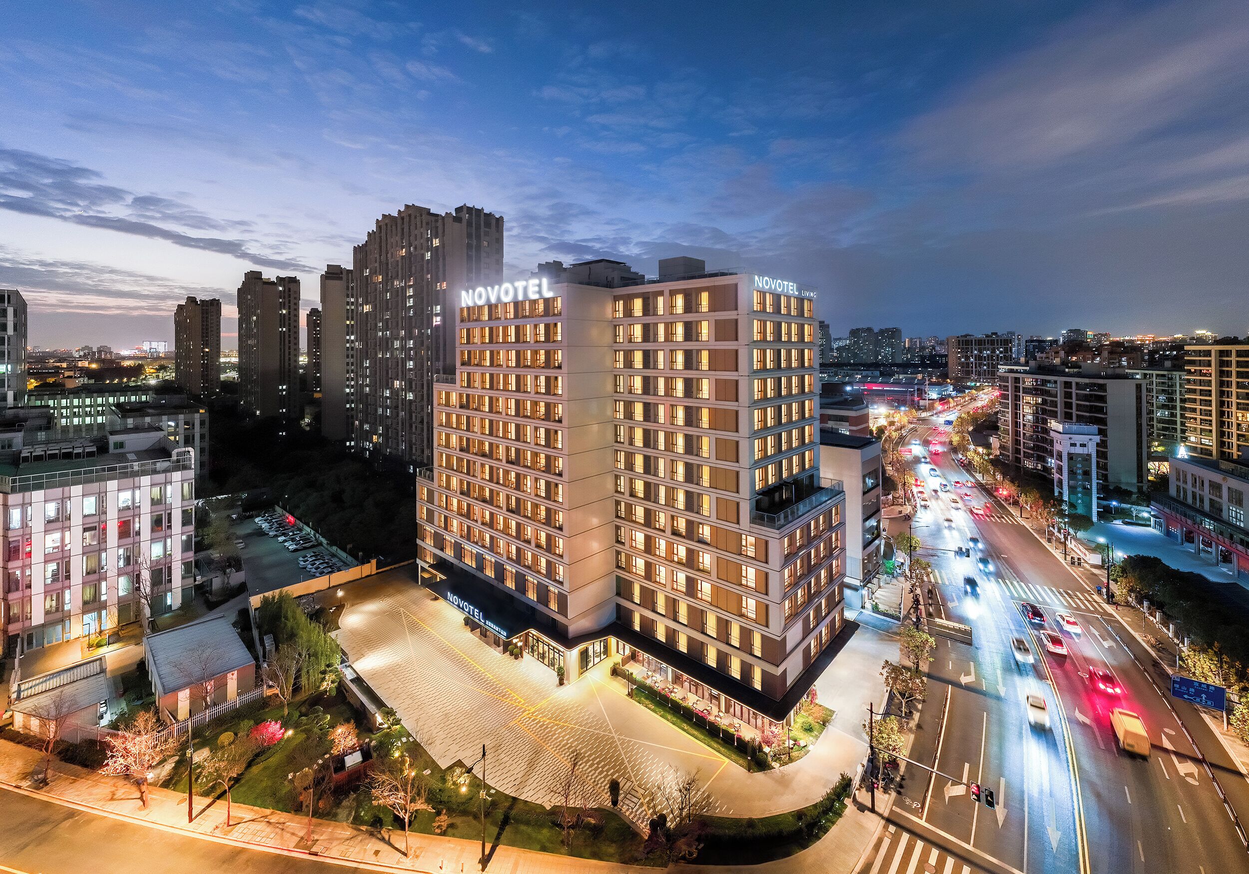 Um hotel Novotel com vários andares, área de estacionamento e vista para o horizonte urbano ao entardecer.