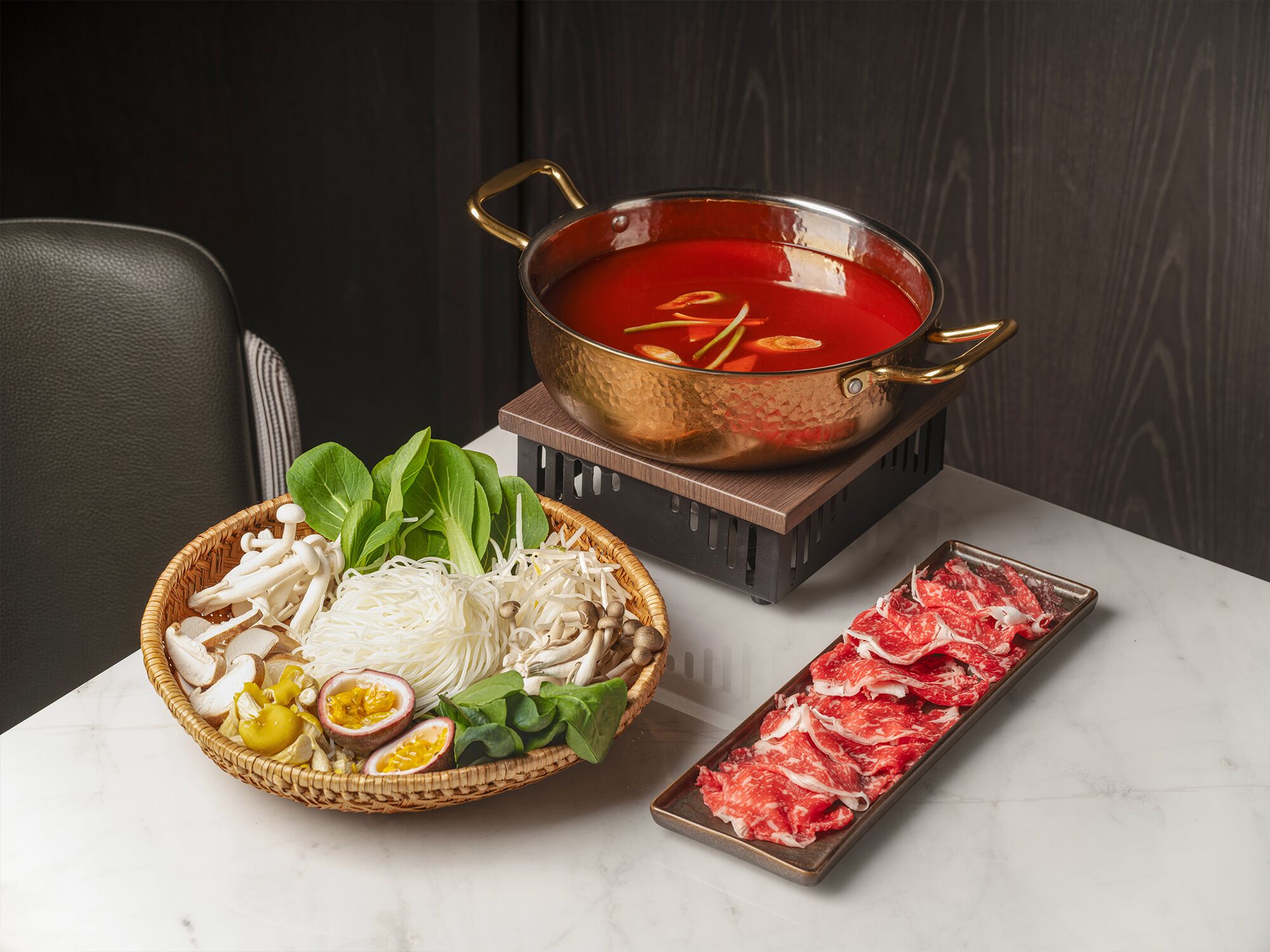Una preparación de olla caliente con un caldo rojo, una bandeja de carne cruda y una cesta de verduras y hongos.