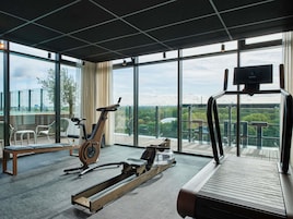 A modern gym with a rowing machine, elliptical, and treadmill, large windows with a city view, and a balcony with outdoor seating.