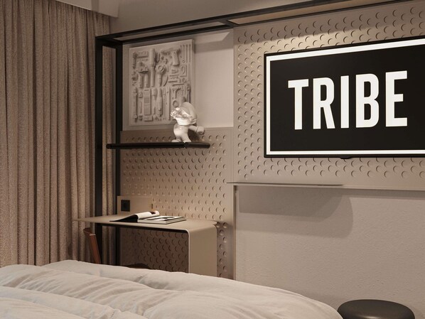 Ein Hotelzimmer mit einem Bett, einem Schreibtisch mit Stuhl, einem Wandregal mit einer Skulptur und einem Schild mit der Aufschrift 'TRIBE'.