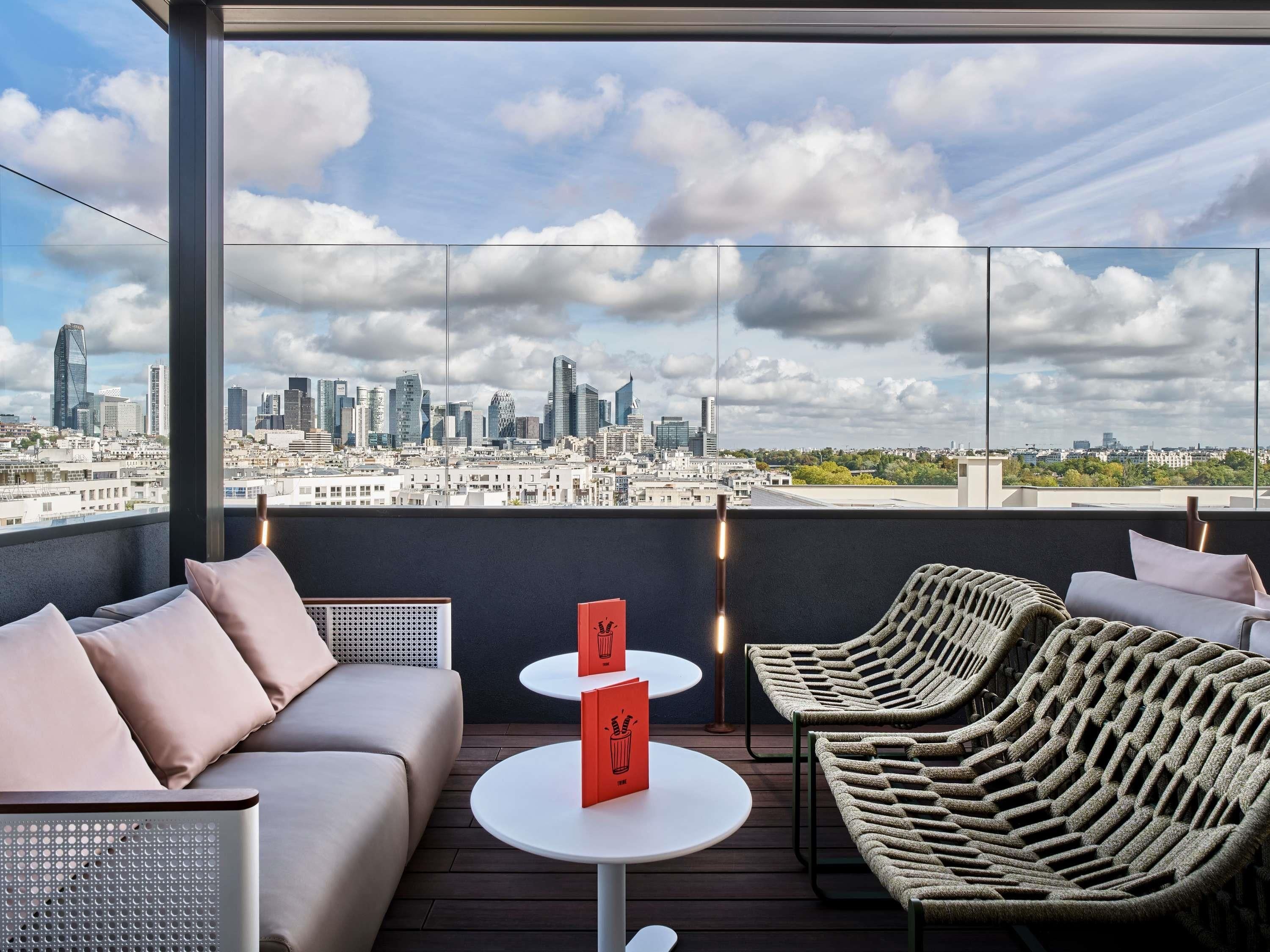 A rooftop terrace with a city skyline view, a sofa, two chairs, a small table, and a red book.