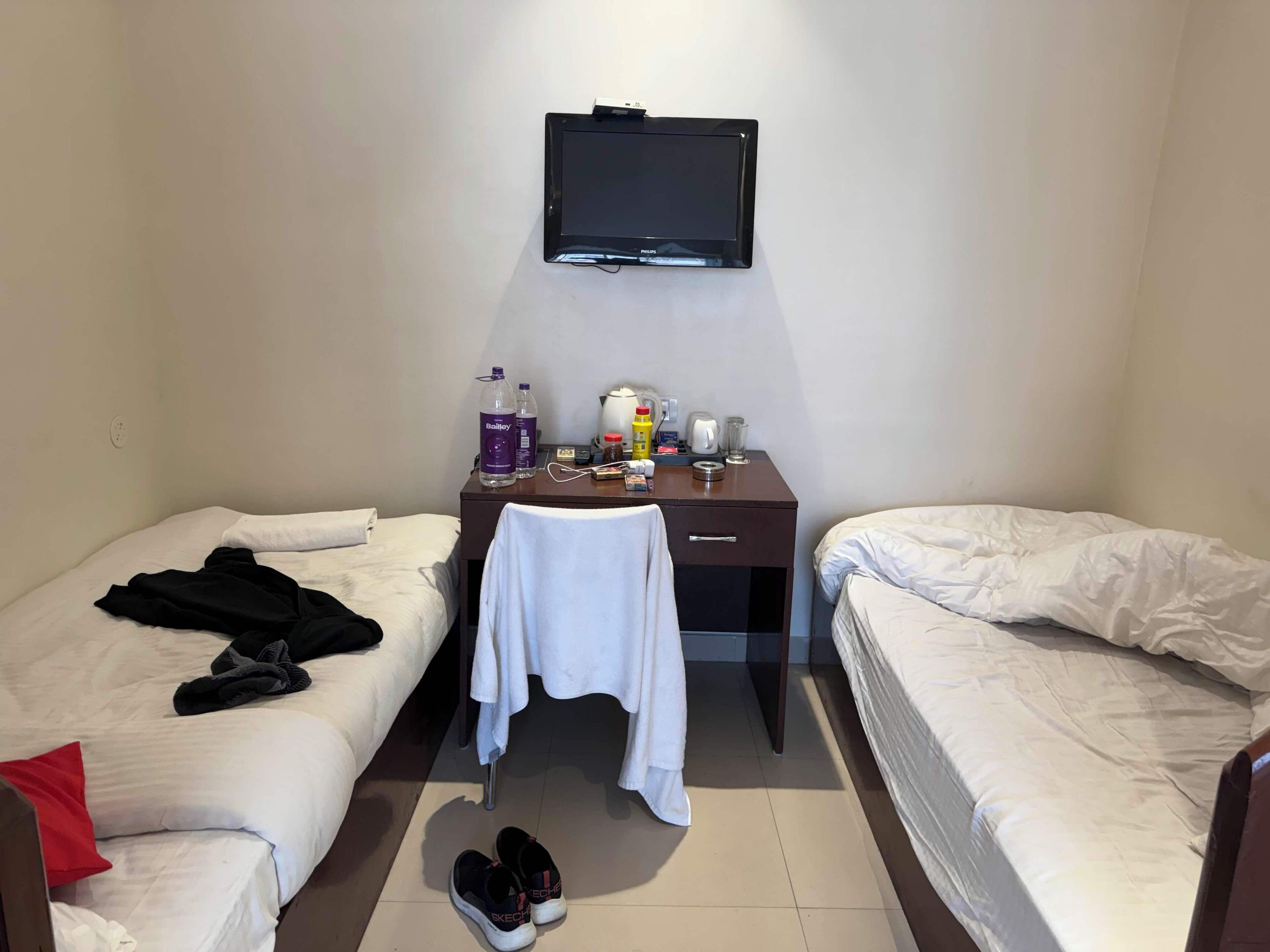 A hotel room with two single beds, a desk with a TV, a chair, a table with toiletries, and a pair of shoes on the floor.