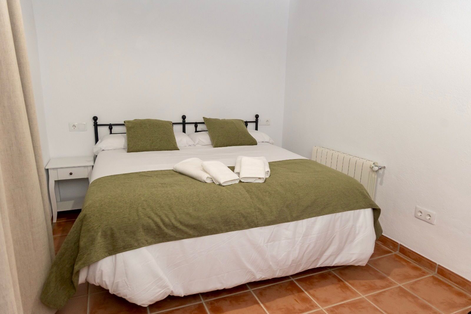 A single bed with a green blanket and pillows, a white duvet, and neatly folded towels on the bed.
