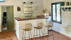 A kitchen with a wooden ceiling, a white wall with a mounted deer head, a bar counter with bar stools, and a window with a view. - Casa Rural Cerro Caña (Belmez)