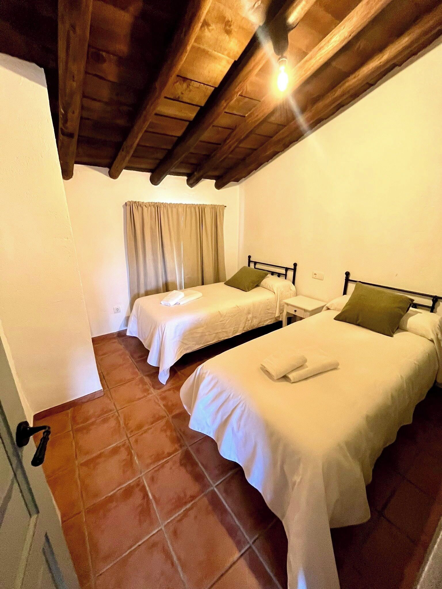 A room with two single beds, a wooden ceiling, a window with curtains, and a tiled floor.