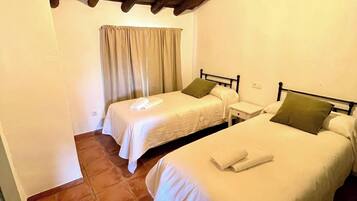 A room with two single beds, a wooden ceiling, a window with curtains, and a tiled floor.
