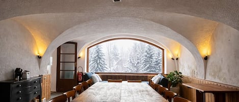 A dining area with a long wooden table, chairs, and a view of snow-covered trees through a large arched window.