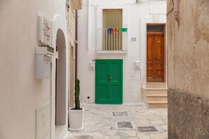 Un stretto vicolo con una porta verde con persiana, una pianta in vaso e una porta di legno in fondo.