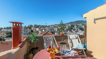 Un balcon avec une table dressée pour un repas, une bouteille de vin et une assiette de fruits. Le balcon offre une vue sur le paysage urbain, avec des bâtiments et une montagne à l’horizon.