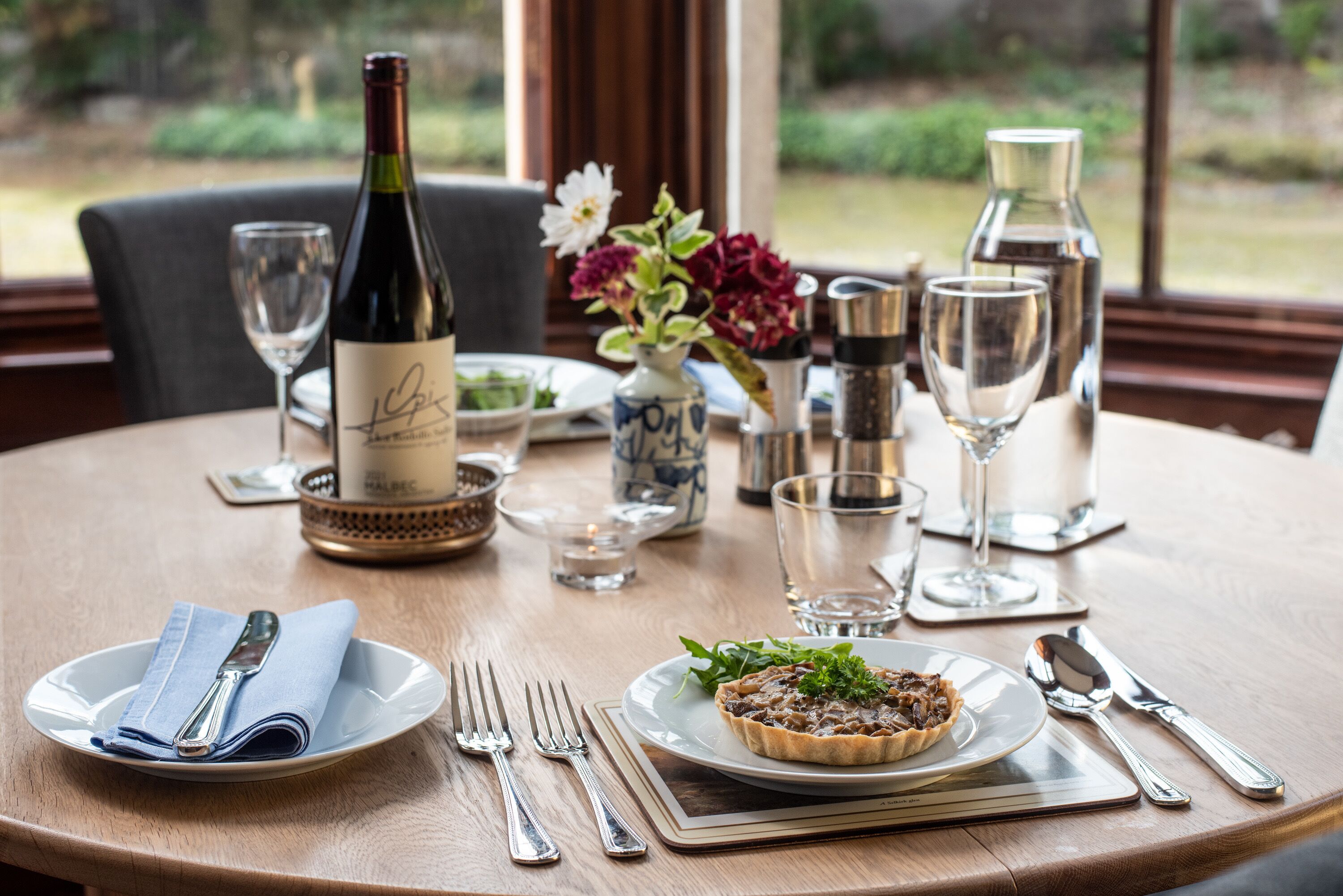 A dining table set with plates, cutlery, a wine bottle, and a vase with flowers.