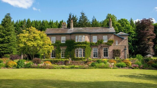 Un'ampia casa a due piani con facciata simmetrica, numerosi camini e un giardino curato con arbusti e alberi diversi.
