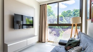 Un salon moderne avec un téléviseur à écran plat fixé au mur, un canapé avec des coussins, une lampe sur pied et une grande fenêtre donnant sur un paysage verdoyant.