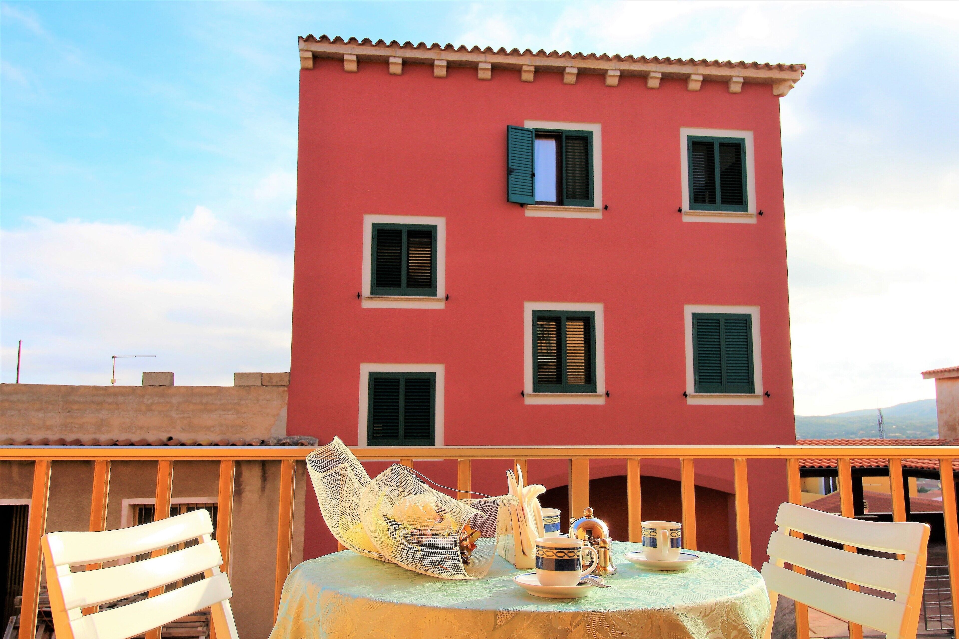 Ein Balkon mit einem Tisch für zwei Personen, einem Stuhl und Blick auf ein rotes Gebäude mit zahlreichen Fenstern und Fensterläden.