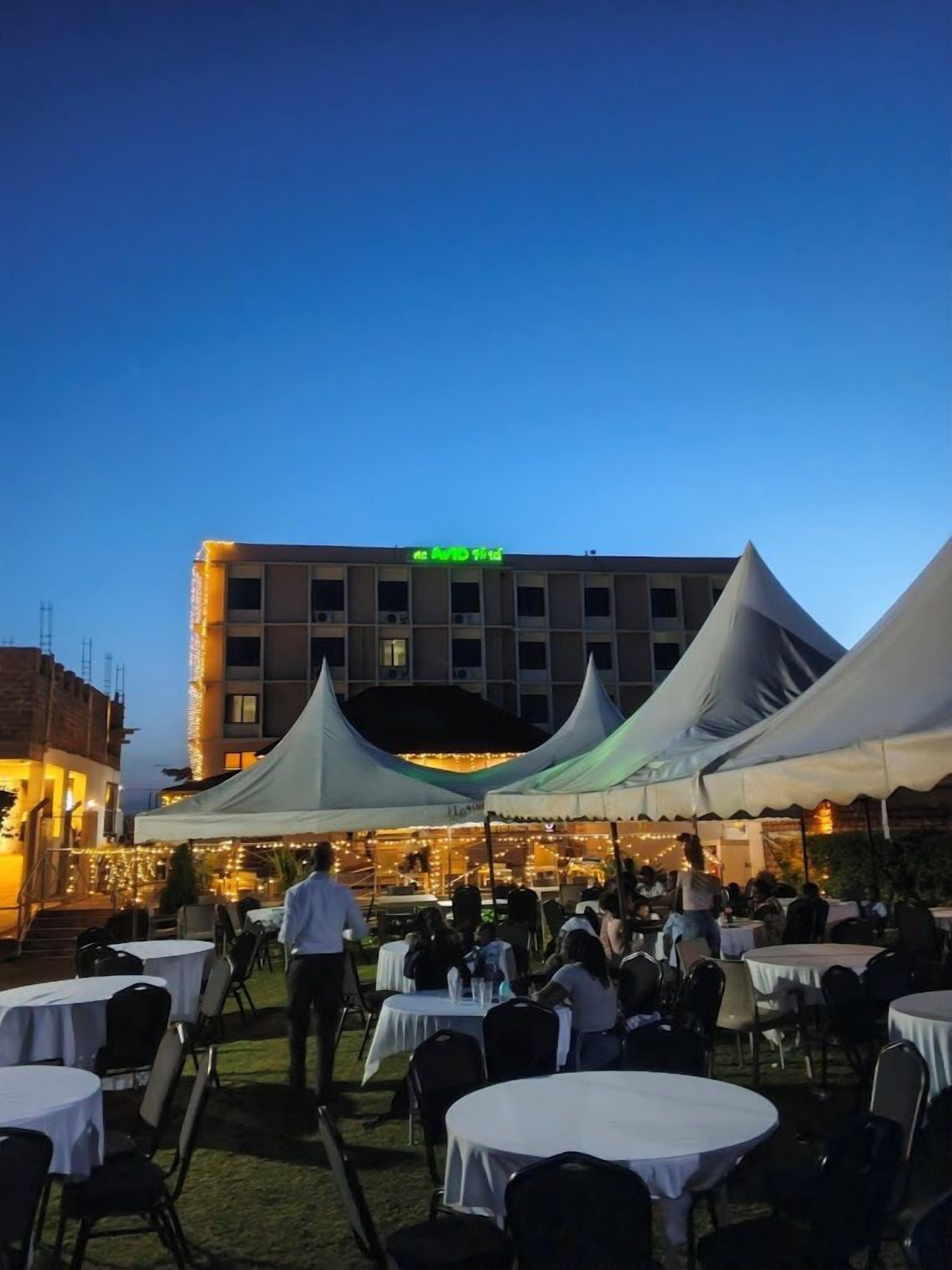 Un espacio de comedor al aire libre con mesas y sillas redondas, un edificio con un letrero al fondo y toldos blancos que ofrecen sombra.