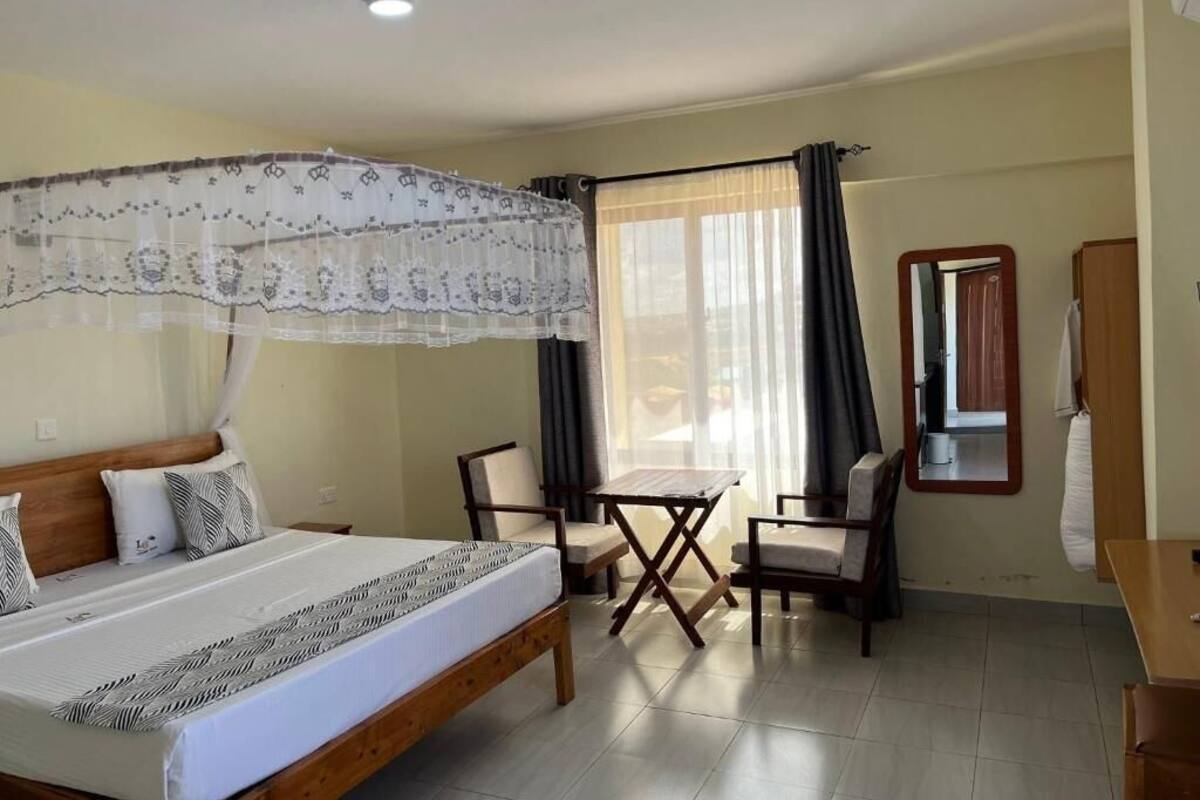A room with a bed, a small table, two chairs, a window with curtains, and a mirror on the wall.