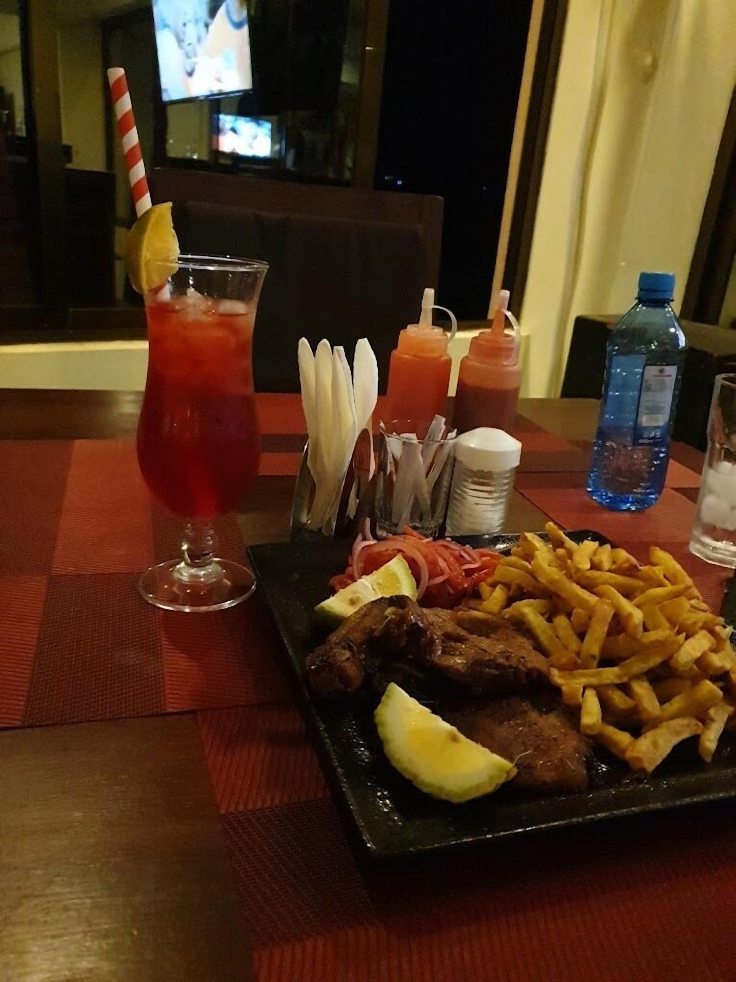Een eettafel met een maaltijd bestaande uit gegrild vlees, friet en een rood drankje met een citroen.